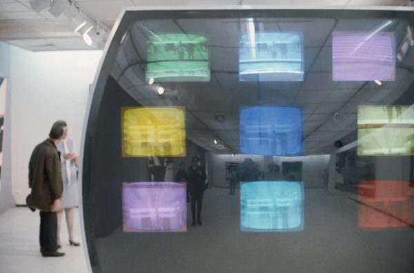A large TV screen with a three by three grid of smaller TV screens inside tinted in different colors. Behind this a person stands looking at something in a gallery space.