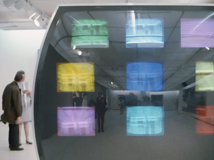 A large TV screen with a three by three grid of smaller TV screens inside tinted in different colors. Behind this a person stands looking at something in a gallery space.