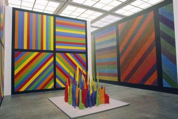 A tall gallery space with floor to ceiling artworks filled with colorful stripes in black square outlines, some are horizontal, some diagonal, and some are vertical. In the middle of the room is a group of colorful shards standing upright.