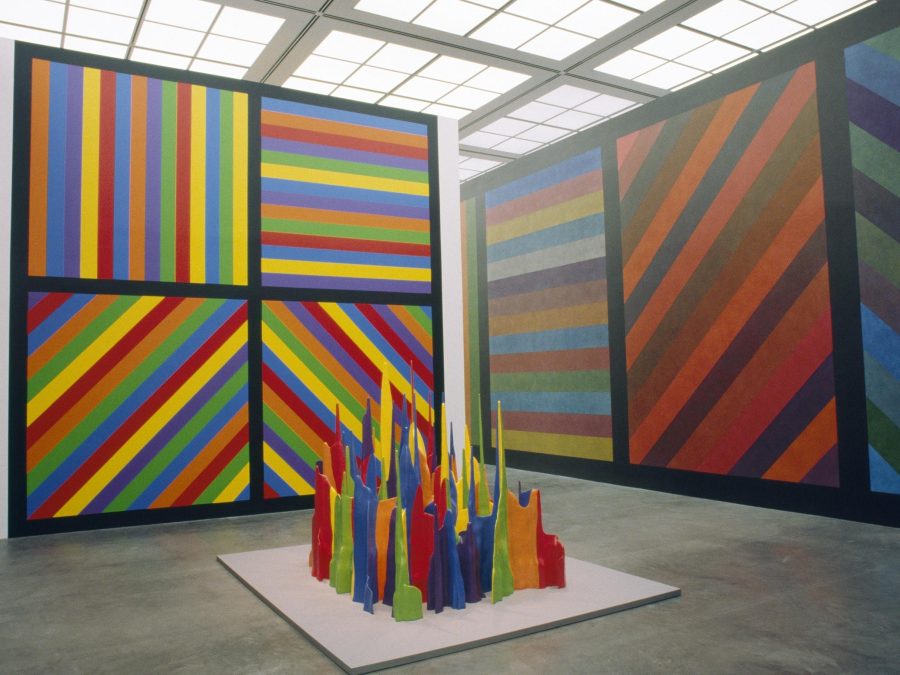 A tall gallery space with floor to ceiling artworks filled with colorful stripes in black square outlines, some are horizontal, some diagonal, and some are vertical. In the middle of the room is a group of colorful shards standing upright.