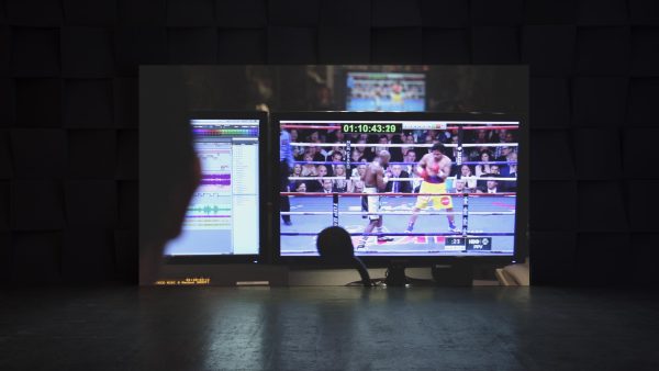 Projected on the wall of a dark room is the image of two monitors, one playing a boxing match, the other showing multiple bars of sound waves.