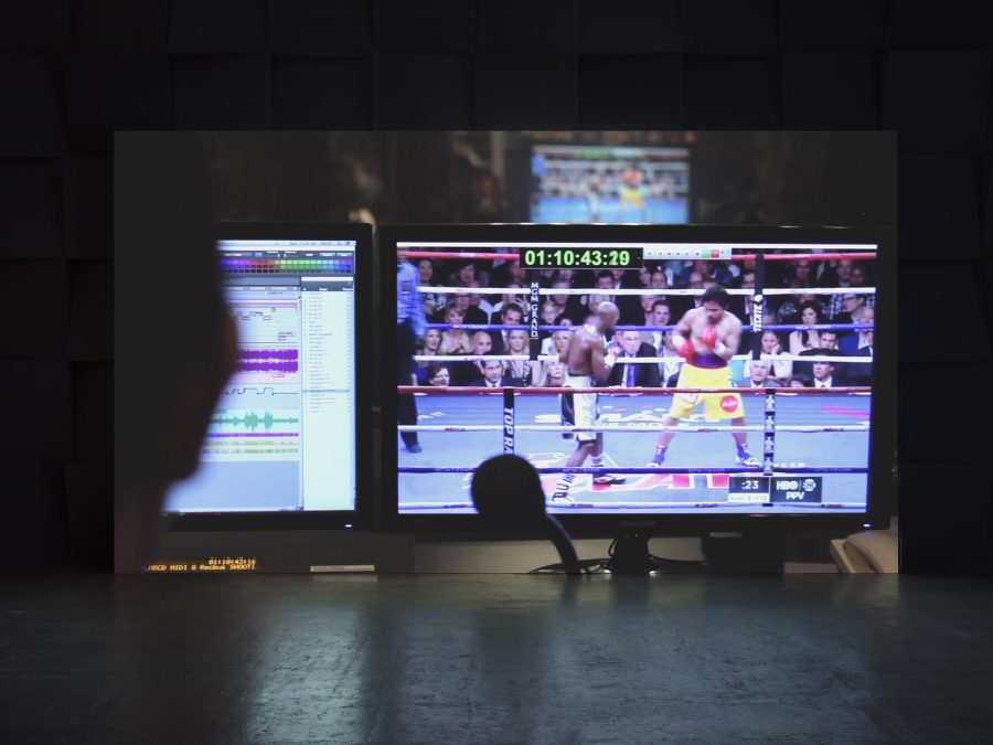 Projected on the wall of a dark room is the image of two monitors, one playing a boxing match, the other showing multiple bars of sound waves.