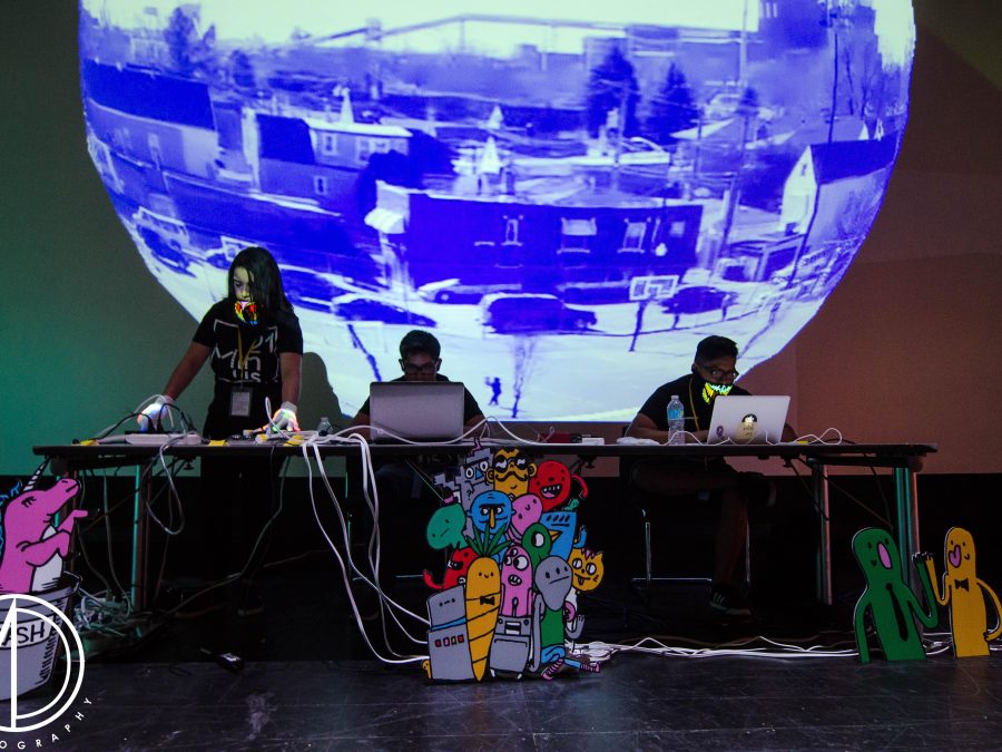 A projected sphere of a warped city landscape is foregrounded by three people at tables with laptops and electronics.