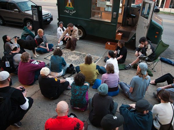 A group of people sitting around a rug on the sidewalk in front of a foodtruck