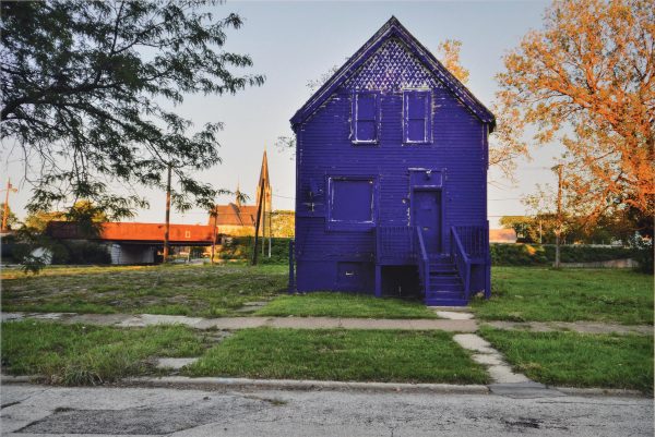 A two-story boarded up house is painted entirely purplish-blue. It is foregrounded by a patchy green lawn, old sidewalk, and damaged road.