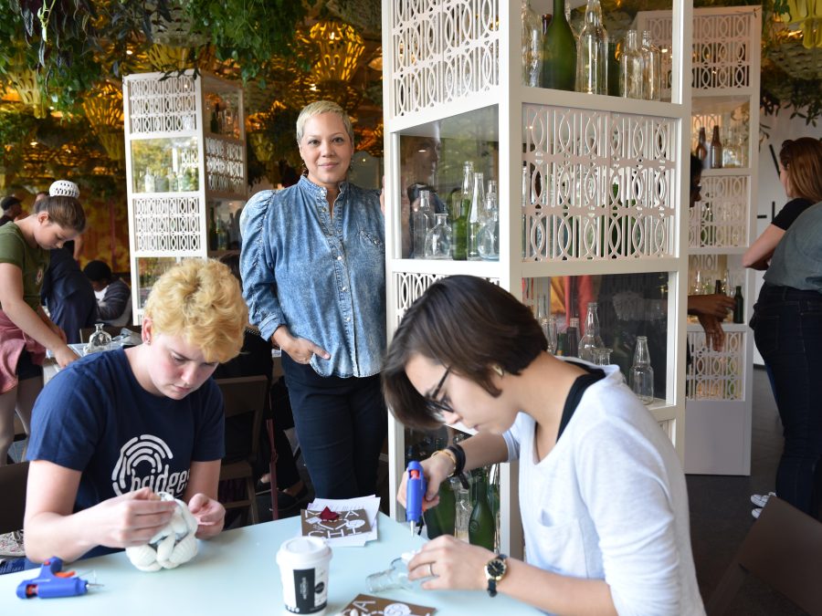 A woman poses next to a white shelving unit filled with glass bottles in a room filled with people working on art projects at tables