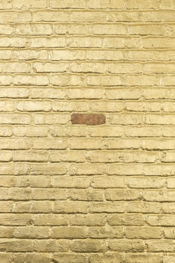 Closeup of a brick wall mostly covered in gold with one brick in the middle left in its natural state