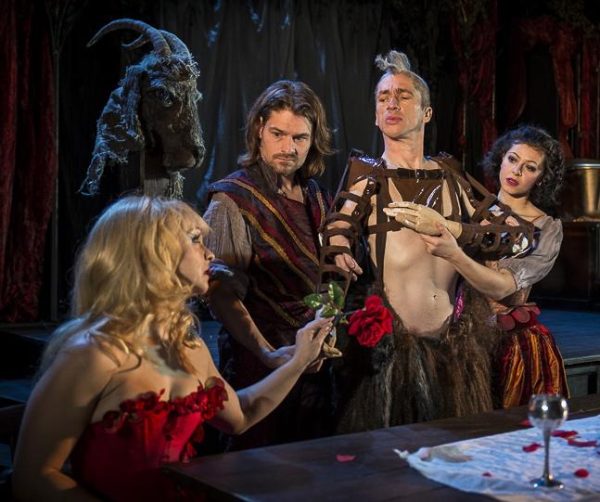 Two people help a man in furry pants with his arms as he hands a rose to a blonde woman dressed in red as a fake ram's head looks on