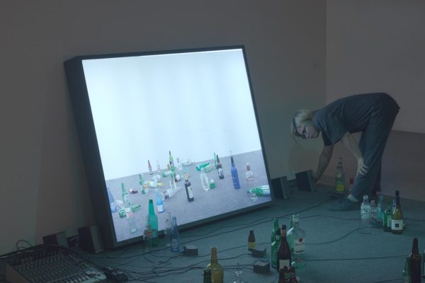 A person bends down to adjust a speaker next to a screen displaying a variety of bottles sitting or laying on a floor. The screen leans against the wall of a darkened room filled with liquor bottles and a sound system.