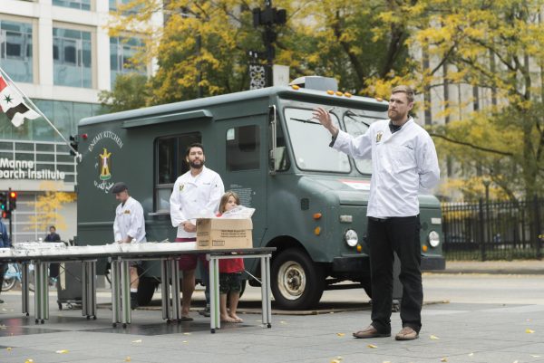 A male-presenting person in a white chef coat gestures as if speaking while two others look on from behind a table next to the Enemy Kitchen food truck