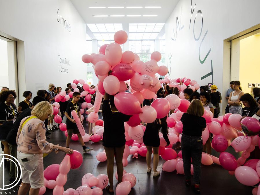 A young person's back is to us as they lift a cluster of pink balloons over their head in the MCA's atrium. Surrounding them are a sea of balloons and people doing similar playful motions.