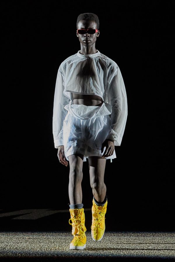 A dark-skinned model walks a fashion runway wearing narrow red sunglasses, a sheer white long-sleeved top and shorts set, and tall bright yellow boots.