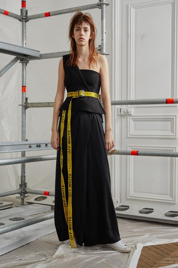 A pale young woman wears a single-shoulder, floor-length black dress cinched with an industrial-yellow belt with black lettering that reads 