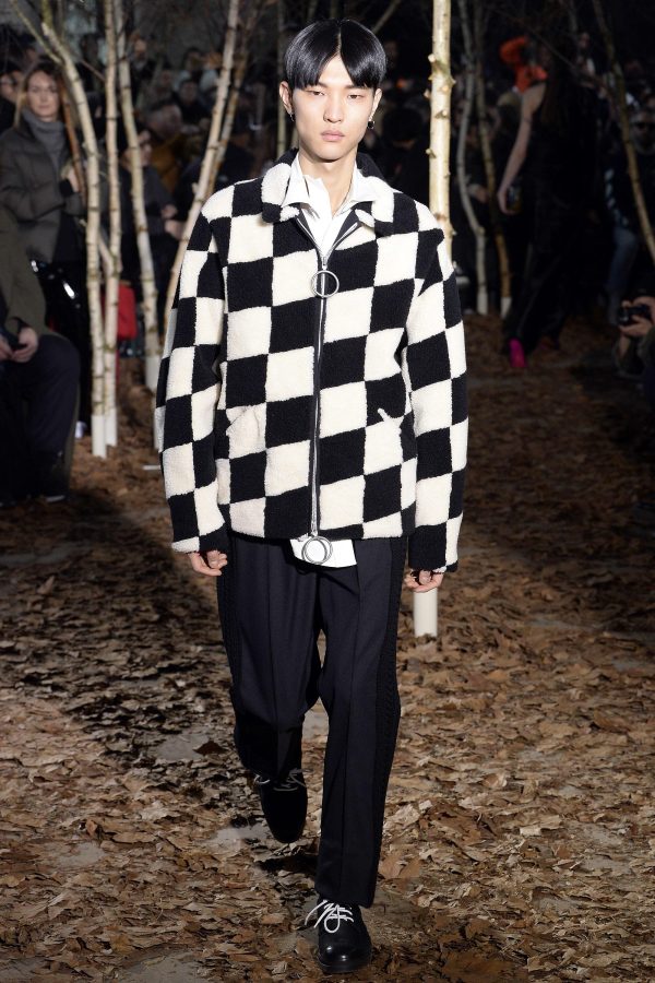 A model walks down a forest-like runway while wearing a boldly checkered black-and-white sherpa fleece jacket and black trousers.