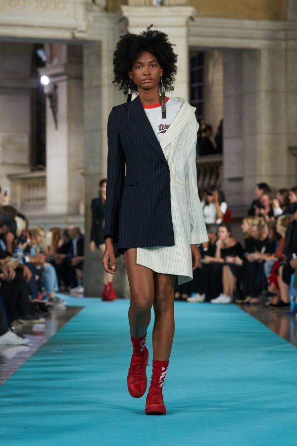 A model walks down a runway while wearing long earrings, an asymmetrical pinstriped suit dress, and red sneakers.