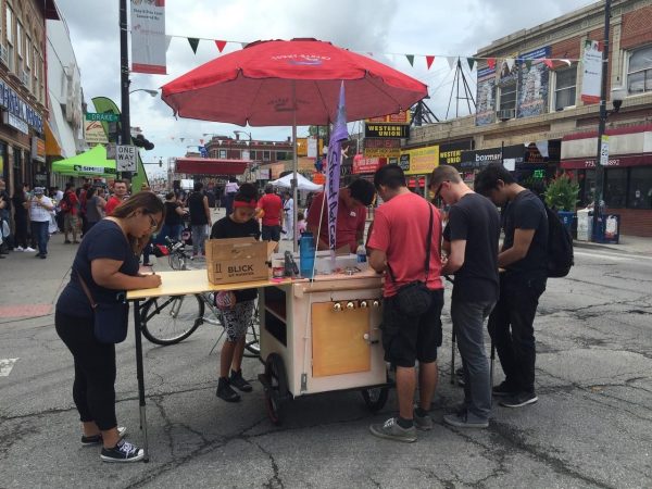 A group of people write and draw using supplies from a wooden cart with an umbrella sticking out of the middle.