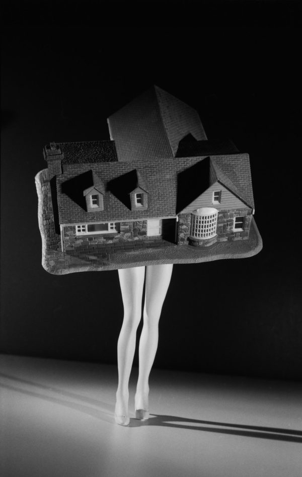 A black and white photograph depicts a model house sitting atop naked, feminine legs wearing nude heels.