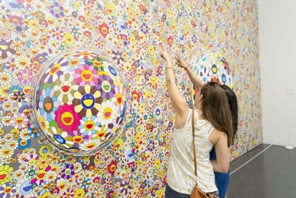Two people point up at a mural of hundreds of Takashi Murakami daisies between two bubble windows that warp the daisies