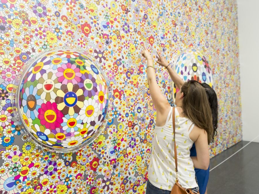 Two people point up at a mural of hundreds of Takashi Murakami daisies between two bubble windows that warp the daisies