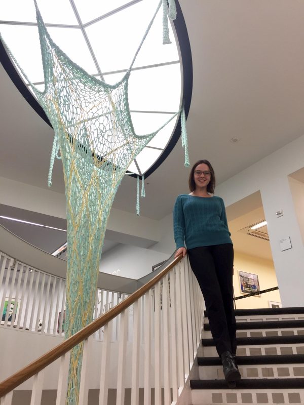 A person poses on a staircase next to a green knit object that extends from the ceiling light down through the opening in the staircase