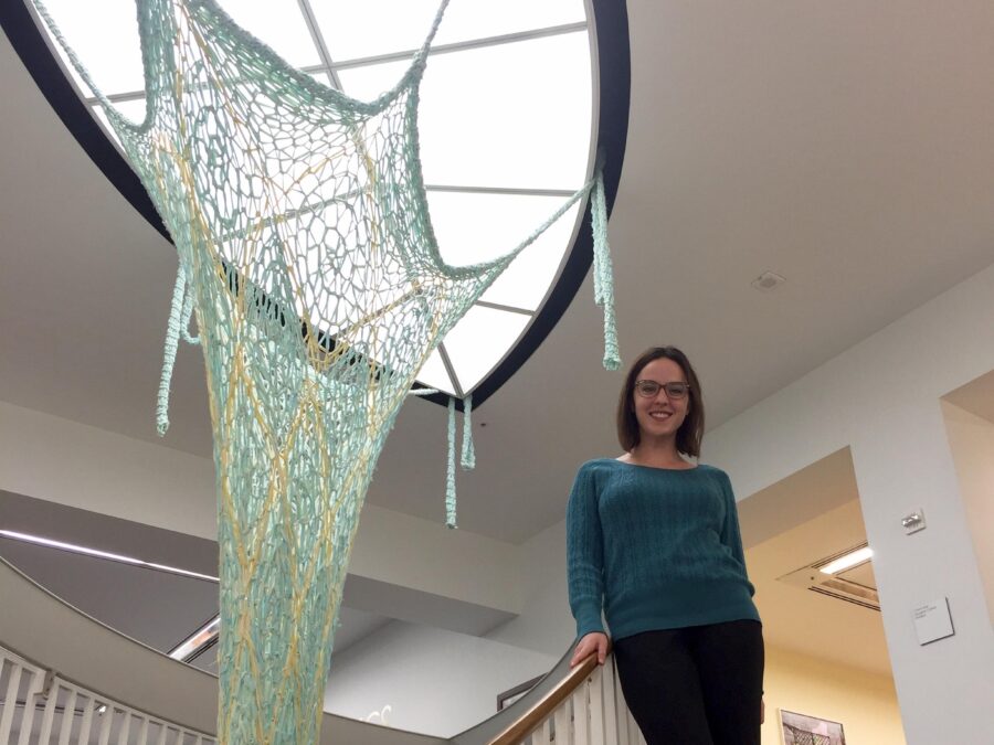 A person poses on a staircase next to a green knit object that extends from the ceiling light down through the opening in the staircase