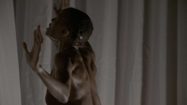With her arms raised, the muscles in her back flexed and straining, her mouth agape, a dark-skinned woman stands looking up at a translucent grey curtain.