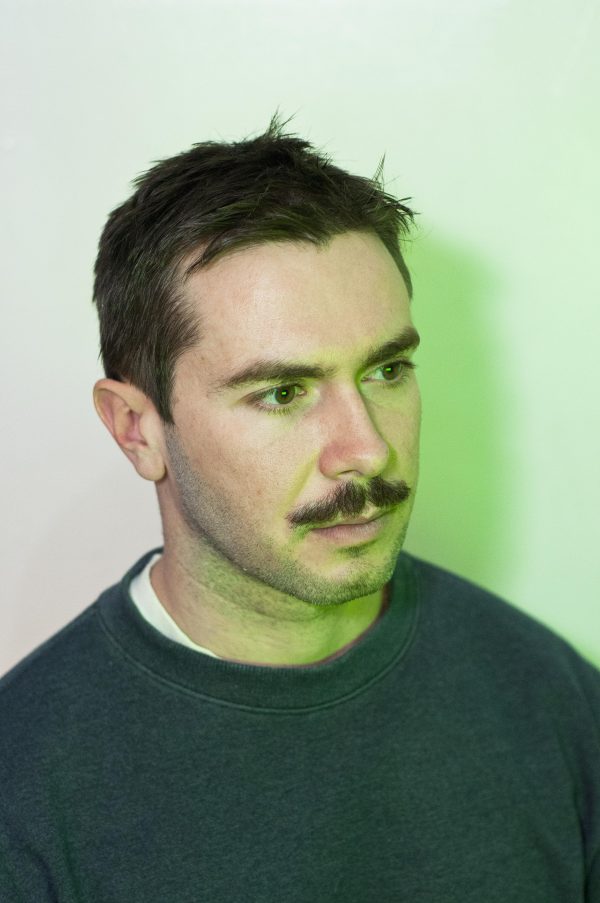 A male-presenting person with light skin, short brown hair, and a mustache lit by a light that casts a green shadow on the wall
