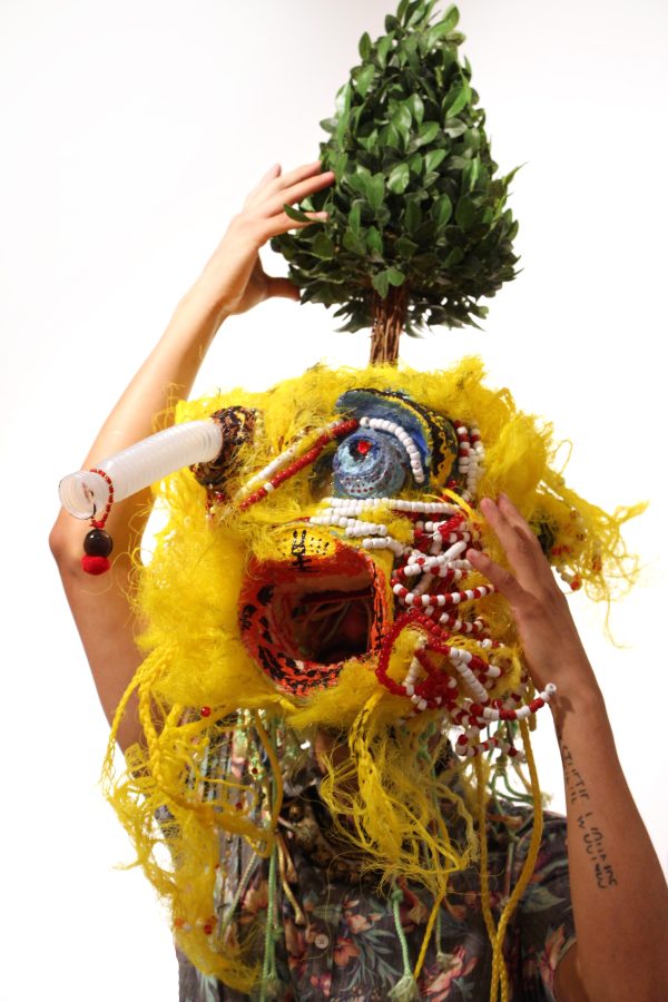 A person in a large yellow mask featuring yarn and a large mouth balances a small tree on their head
