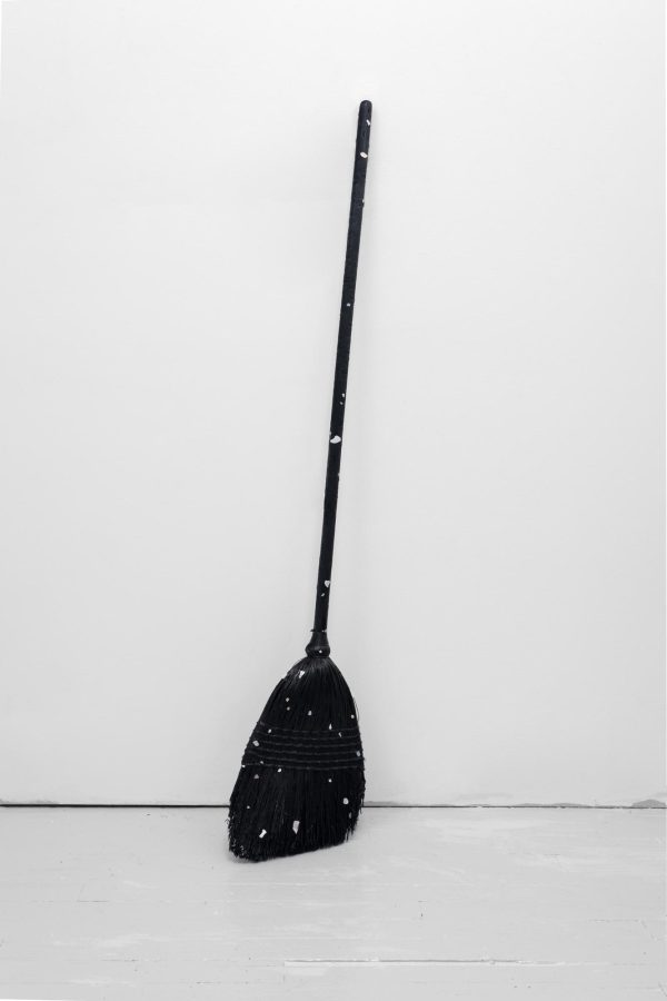 A single charred black broom with white specks leans against a white wall.