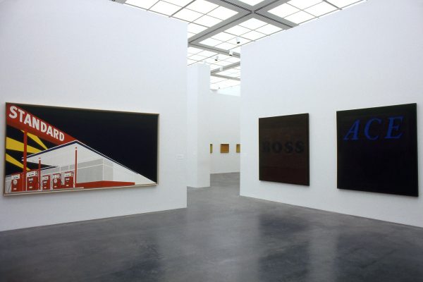 Gallery space with three large paintings hanging on two walls that are divided by a walkway in the middle leading to other rooms with artworks. The artwork on the left is of a Standard gasoline station; the two on the right side are black.