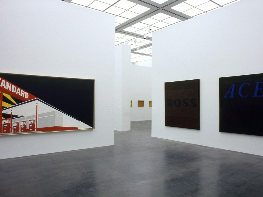 Gallery space with three large paintings hanging on two walls that are divided by a walkway in the middle leading to other rooms with artworks. The artwork on the left is of a Standard gasoline station; the two on the right side are black.