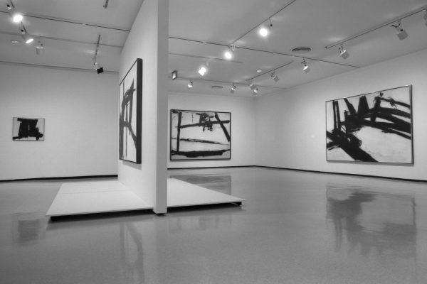 Gallery installation of several large, abstract paintings hung on separate walls with a divider wall in the middle featuring another work
