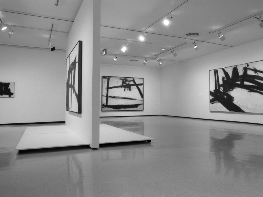 Gallery installation of several large, abstract paintings hung on separate walls with a divider wall in the middle featuring another work