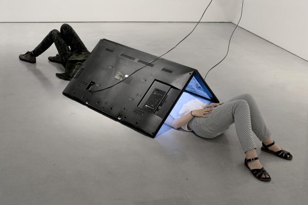 Two people lay on the floor beneath a tent-like structure made from two large digital screens. Their heads are hidden beneath the screens.