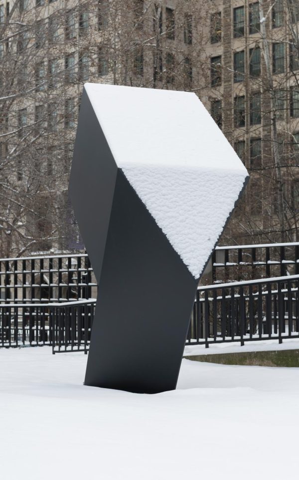 A matte black geometric sculpture juts out from the snowy ground. Two of its faces, one square and one triangular, are perfectly covered by a white layer of snow.