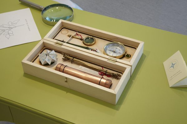 A box made of light wood holds small tools like a magnifying glass, compass, and telescope. It rests on a table alongside a larger magnifying glass and two written materials.