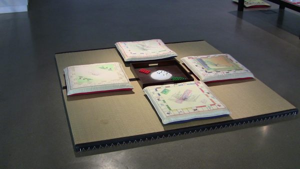 Four square pillows decorated with patterns resembling the board game Monopoly are arranged on a slightly raised platform around a box holding house and hotel playing pieces.