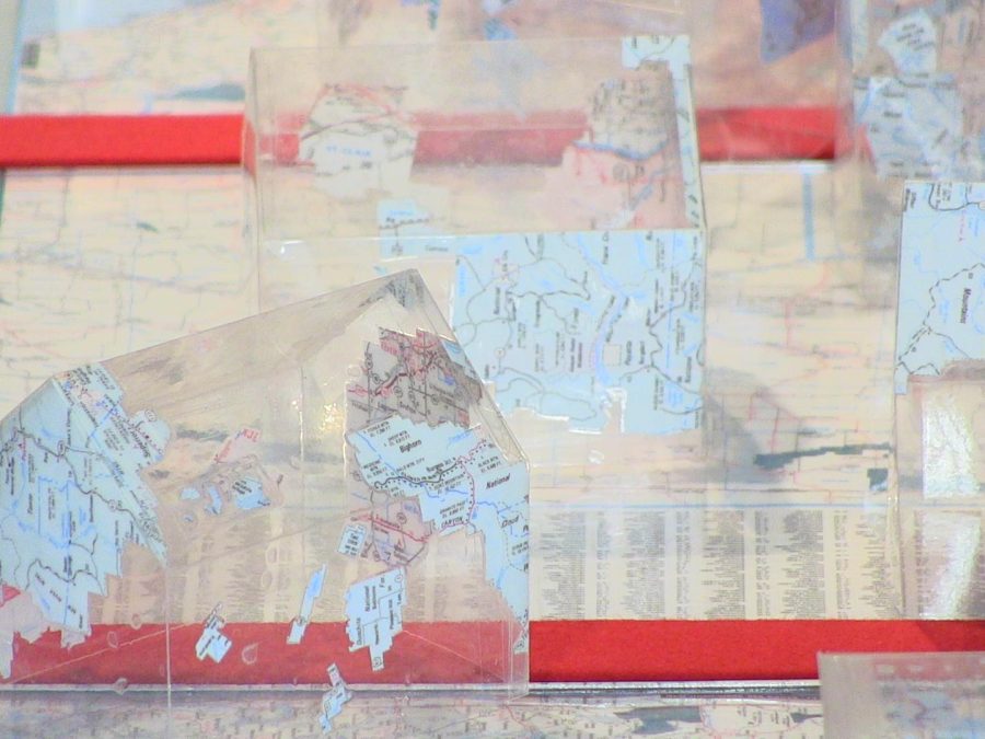 Various translucent maps, assembled into the three-dimensional shape of houses, are arranged on a table covered with more maps.
