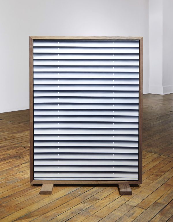 An rectangular, framed artwork that resembles a window shade with the blinds closed in the middle of the room on two wooden blocks