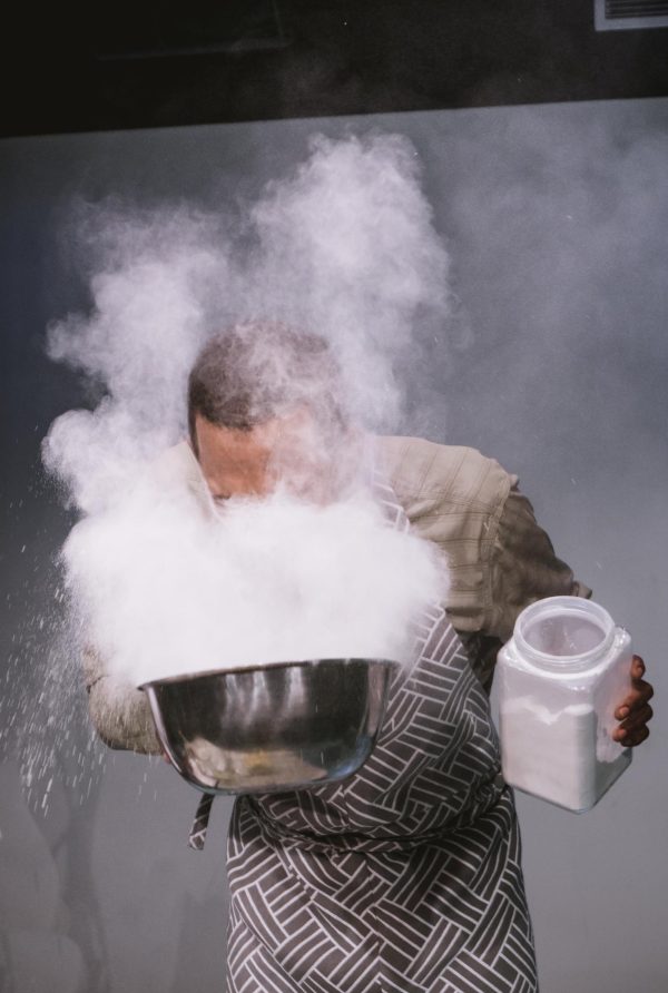 A man bends at the waist to blow into a large metallic bowl full of a white powder. The powder plumes into his face and around his head, obscuring his facial features.
