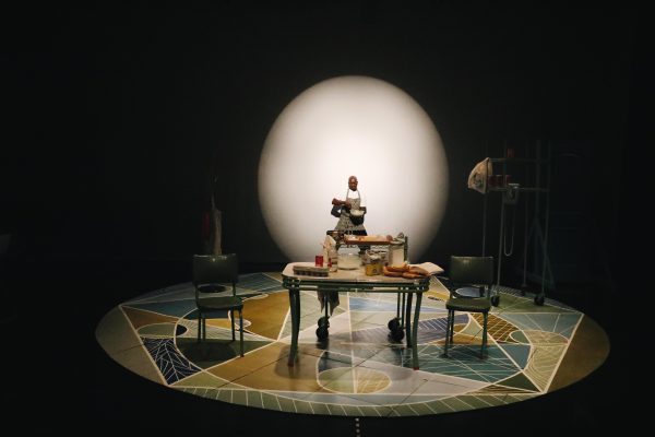 A dark-skinned man stands on a stage set like a kitchen with a mosaic circular floor and a large white sphere looming behind him.