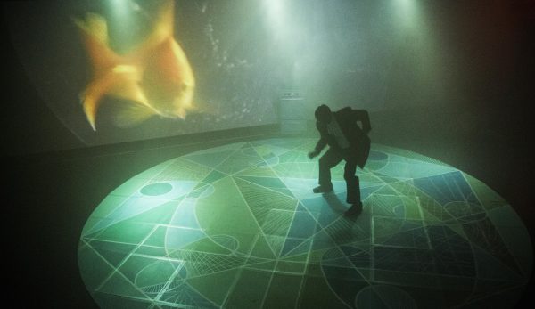 A man in a businesslike suit crouches as if dancing on an illuminated stage. The floor beneath him features an intricate pattern of various greens, while the screen behind him shows a blurry projection of a goldfish.