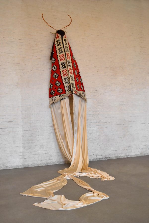 A fabric resembling an ornate rug or cape is mounted on a white brick wall. From the cape's bottom trails long, silky bolts of fabric, and from its top protrudes antennae-like twigs.