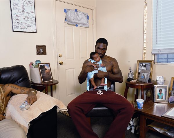 In this photo, a young, dark-skinned man holds a very young baby to his chest in what appears to be the living room of a home.