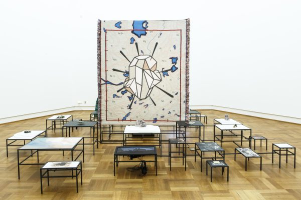 A large tapestry of a gem pierced by needles stands vertically in an art exhibition. It is surrounded by over a dozen metal tables of different sizes and heights.