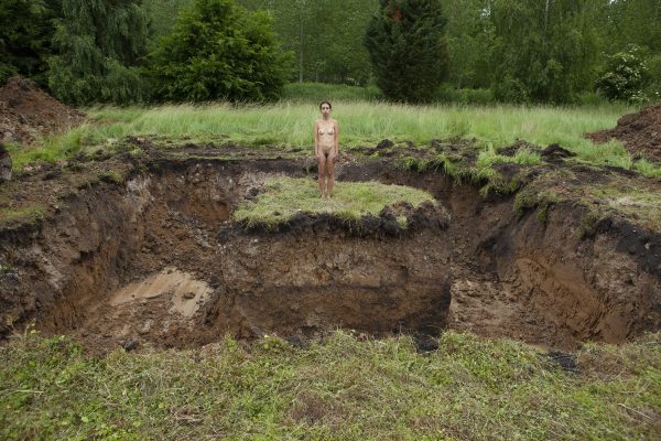 A naked female stands on a grass island surrounded by excavated land