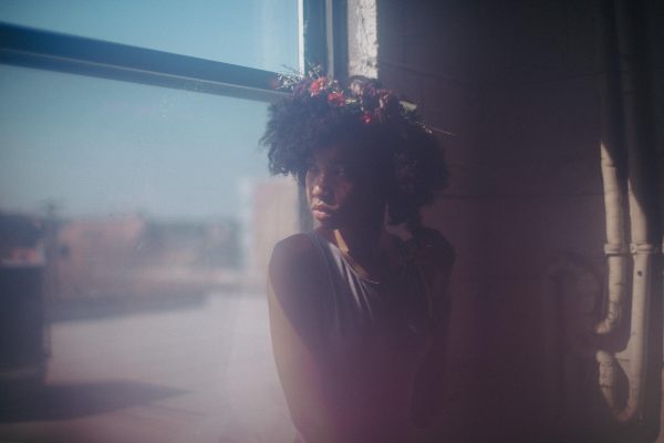 A young black woman, wearing a wreath of flowers in her hair and sitting in a window, turns to look over her shoulder. A misty haze slightly obscures the image.