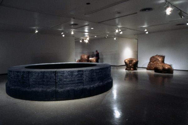 Dimly lit gallery space with a circular black sculpture in the middle of the room and bronze forms behind it