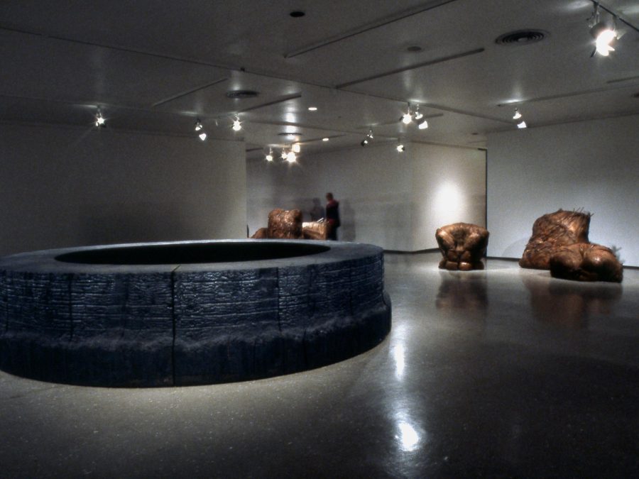 Dimly lit gallery space with a circular black sculpture in the middle of the room and bronze forms behind it