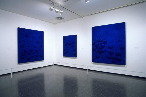 Installation of three blue paintings with objects attached to them in the corner of a gallery